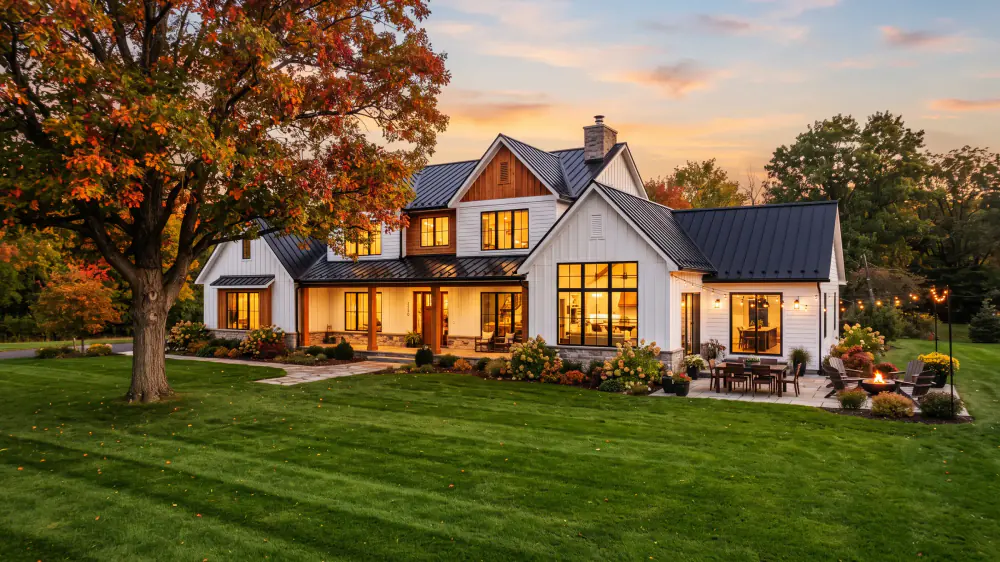 White farmhouse with glowing windows and an autumn maple at dusk
